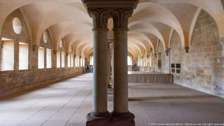 Long stone hall with paired columns, repeated low vaults, and a row of rectangular windows along one wall.
