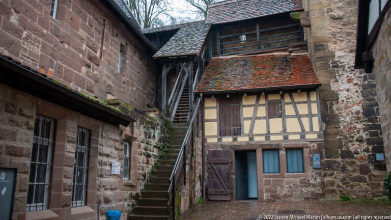 Small half-timbered building and steep exterior stair enclosed by rough stone monastery walls.