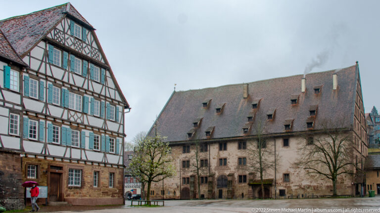 Courtyard between a half-timbered building with shutters and a large steep-roofed stone monastery range.