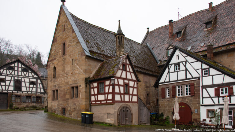 Tall gabled stone building flanked by smaller half-timbered houses along a narrow monastery lane.
