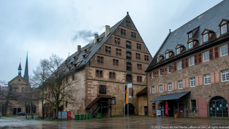 Steep-gabled multi-story building and long stone service range in a paved monastery court, with church beyond.