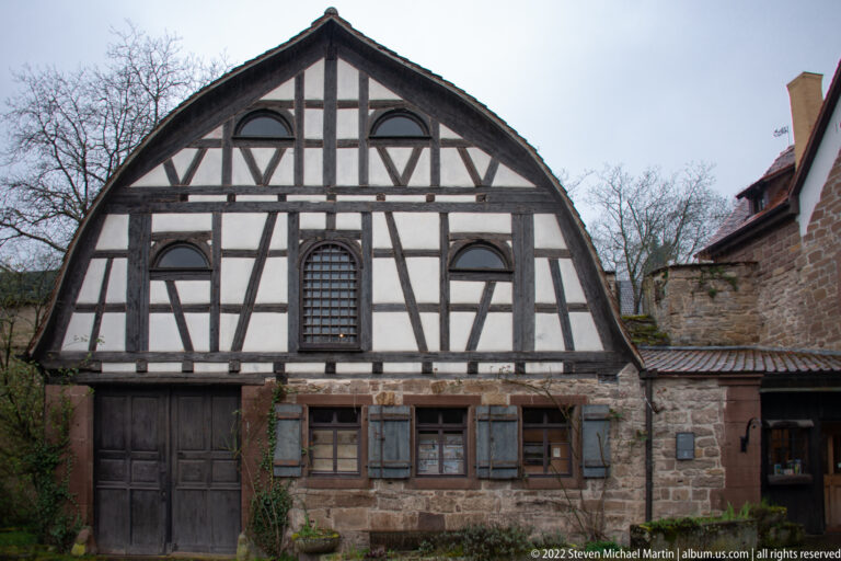 Broad-gabled half-timbered building with pale infill, arched upper openings, shutters, and a stone lower story.