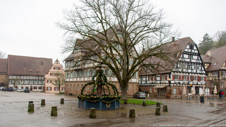 Wet historic square with ornamental fountain, large bare tree, and half-timbered buildings around the perimeter.