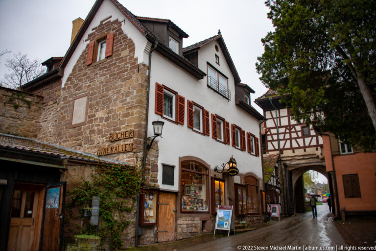 White historic building with red shutters and shopfront beside stone walls and a half-timbered arched passage.
