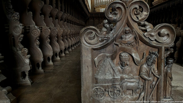 Carved wooden choir-stall end with scrolling foliage and a narrative relief of several figures.