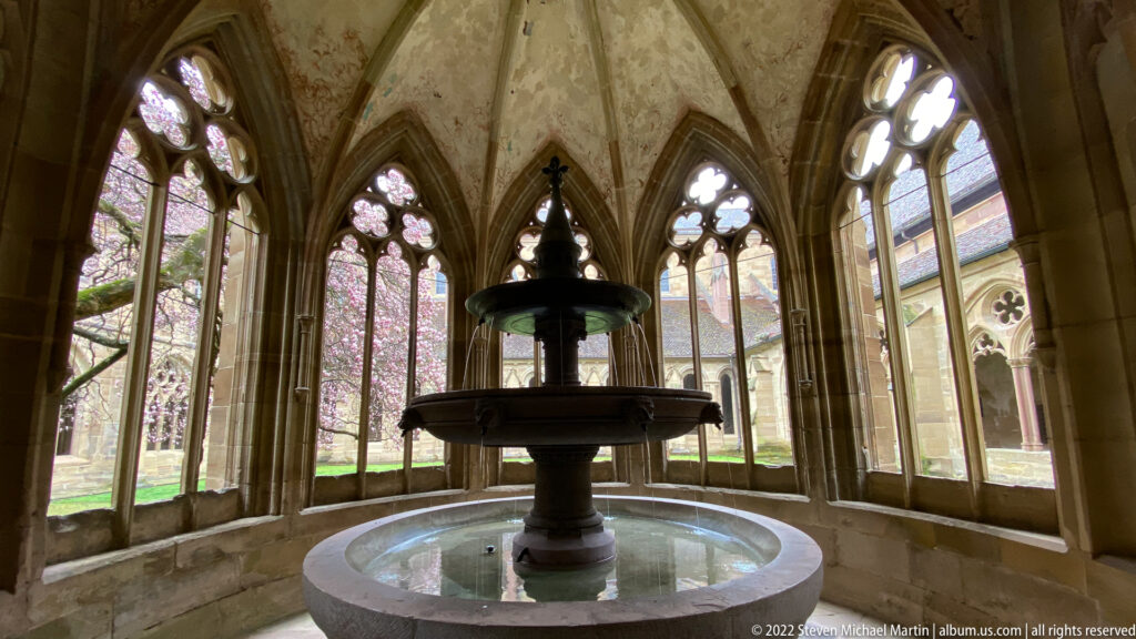 Tiered stone fountain centered before tall Gothic tracery windows overlooking the green cloister courtyard.