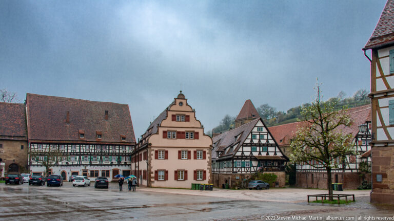 Gabled stucco house with red shutters flanked by half-timbered buildings on a wet square.