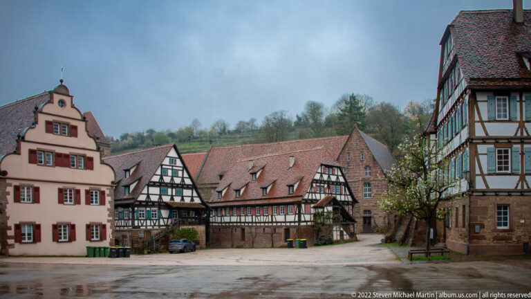 Half-timbered buildings in front of a larger stone range with steep red roof around a paved open space.