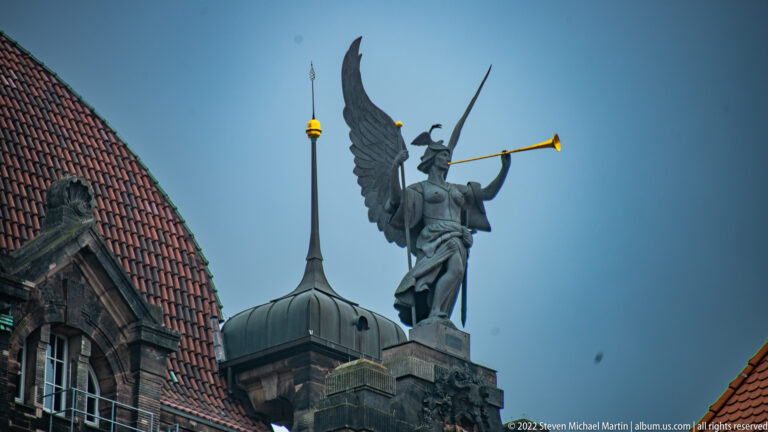 Winged rooftop figure holding a long trumpet beside a dark dome and gilded finial against blue sky.
