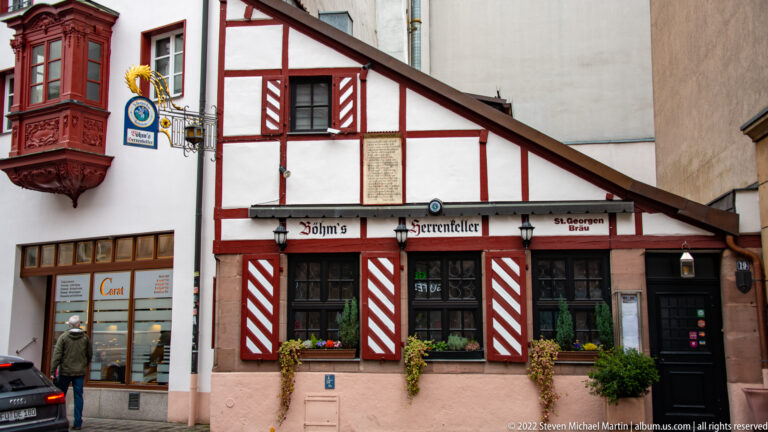 White-and-red half-timbered Böhm’s Herrenkeller façade with striped shutters, signage, and flower boxes.