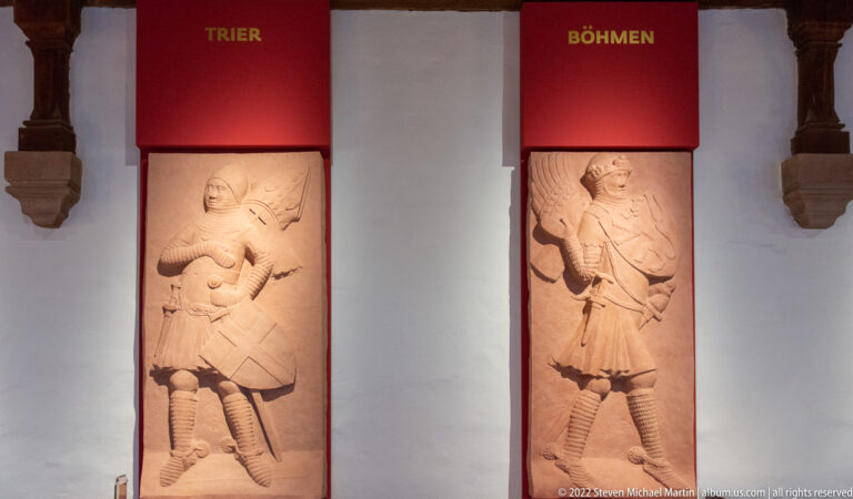 Two carved standing figures beneath red banners labeled Trier and Böhmen in an Imperial Castle exhibition.