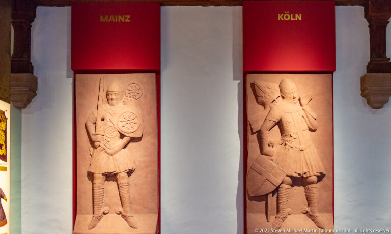 Two carved standing figures beneath red banners labeled Mainz and Köln in an Imperial Castle exhibition.