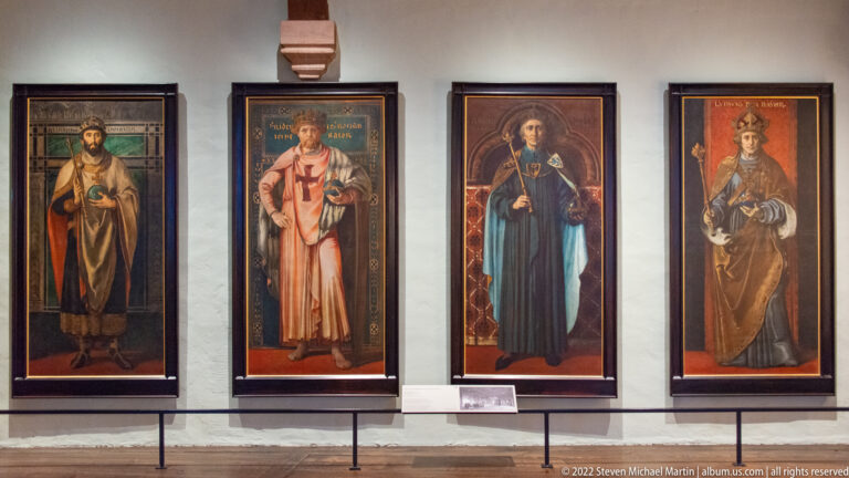 Four narrow framed paintings of standing robed figures displayed in a row on a white wall.