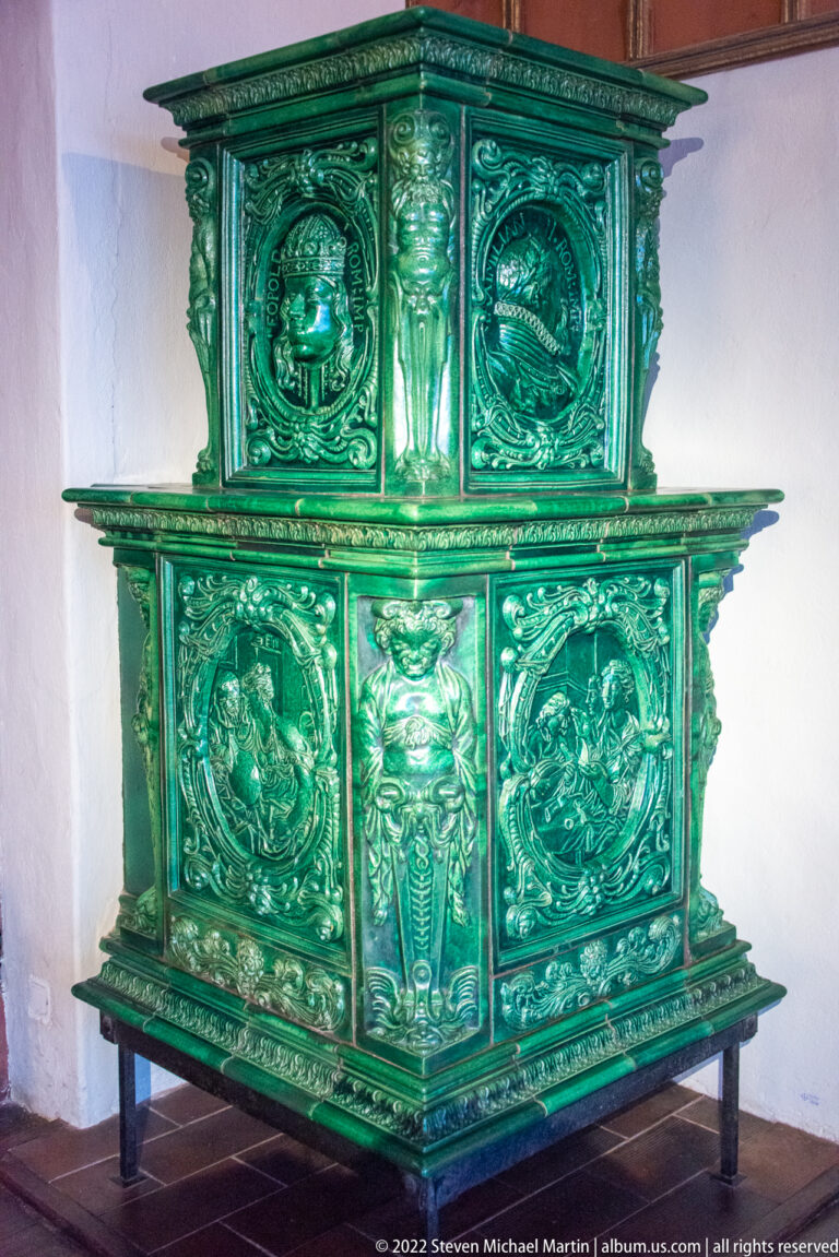 Large green glazed tile stove with relief panels, medallions, figures, and foliage on metal feet.