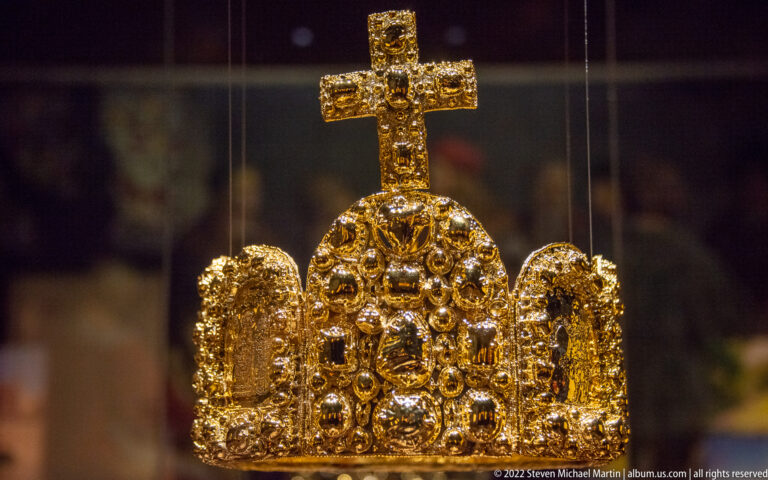 Jeweled gold imperial crown with prominent cross and gem-set arched plates displayed behind glass.