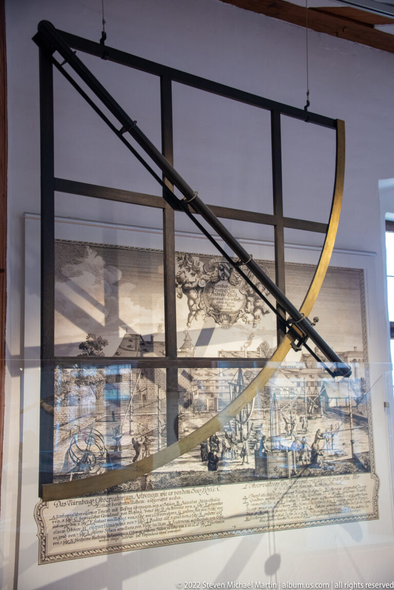 Large historic crossbow mounted horizontally in a museum case with a period illustration behind it.