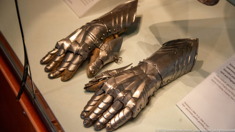 Pair of articulated steel armour gauntlets with segmented fingers and flared cuffs in a museum case.