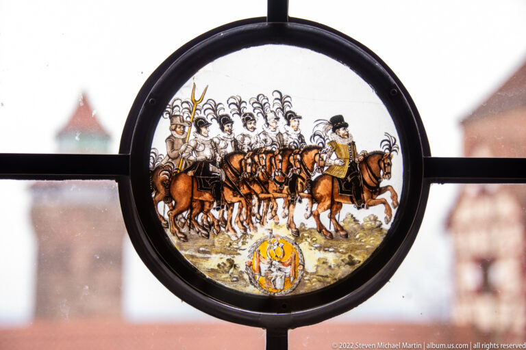 Circular stained-glass panel with a procession of mounted riders and a small heraldic shield below.
