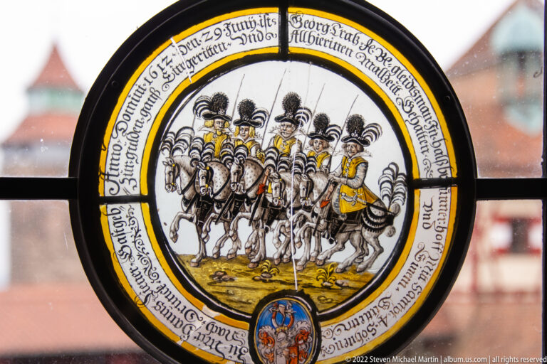 Circular stained-glass roundel with armored lancers, plumed helmets, surrounding inscription, and coat of arms.