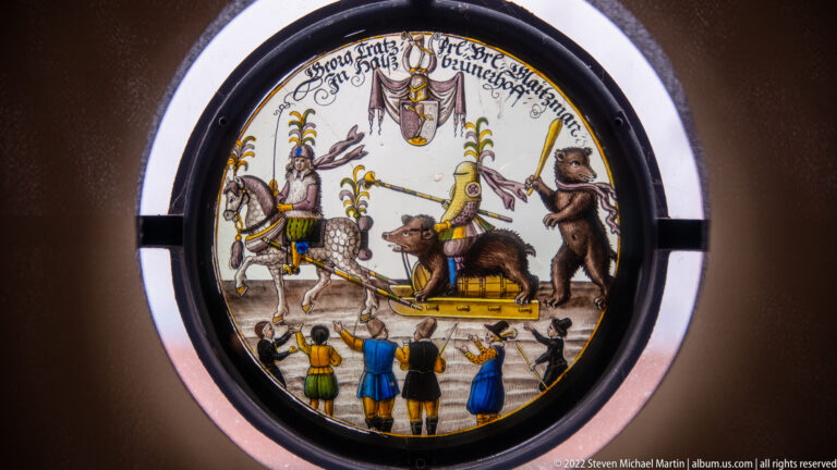 Circular stained-glass panel with armored figures, animal supporters, heraldry, banners, and spectators.