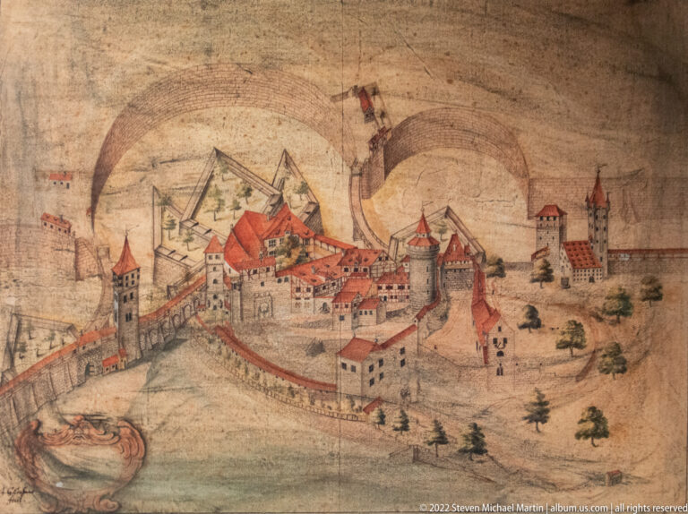 Historic drawing of a fortified castle with red-roofed buildings, perimeter walls, towers, and outer defenses.