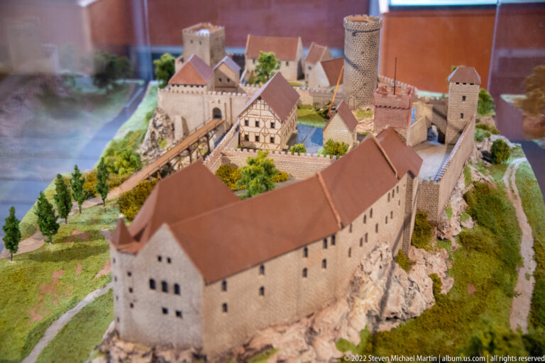 Scale model of a fortified castle with stone walls, towers, timber buildings, bridges, and red roofs.