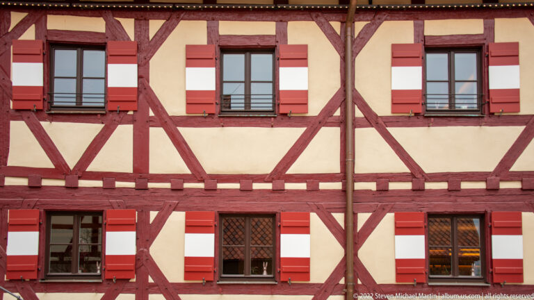 Red timber-framed façade with pale plaster panels, dark windows, and red-and-white shutters.