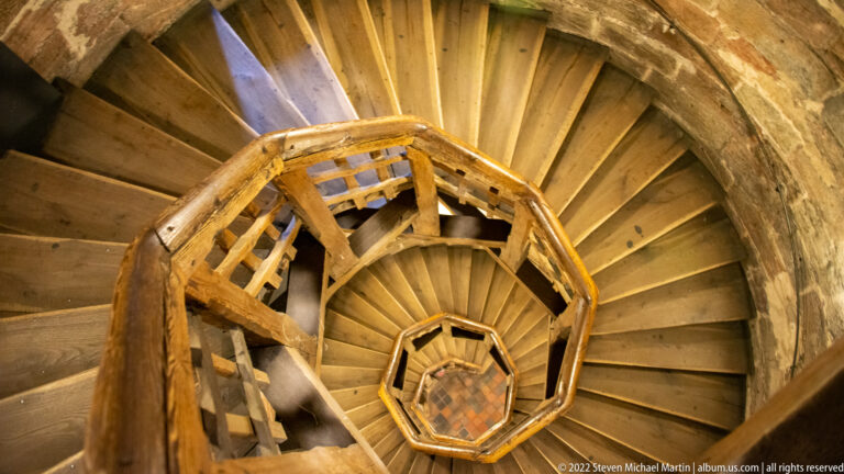 Wooden spiral staircase curling downward inside rough stone walls with a polygonal timber handrail.