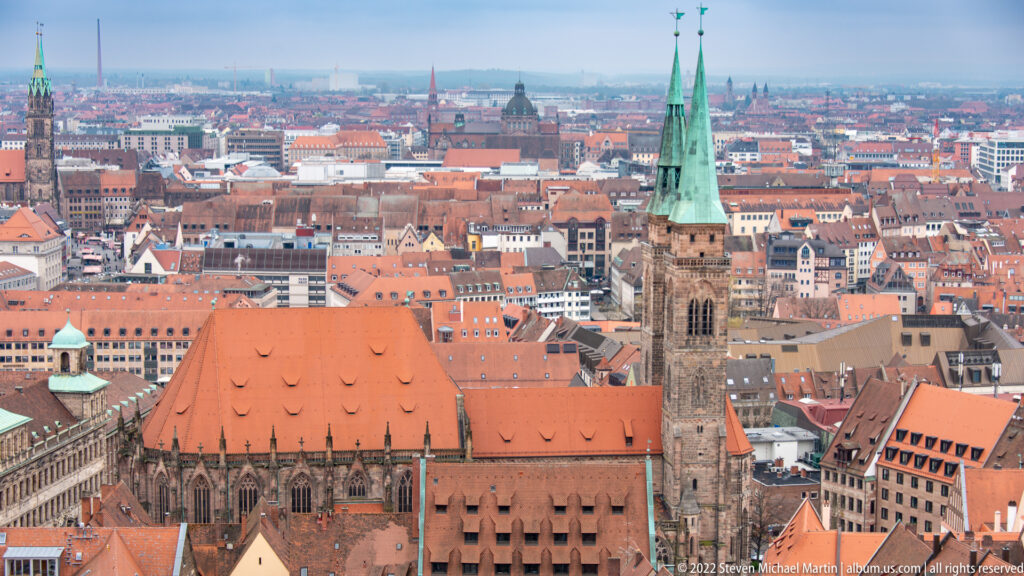 Elevated view of St. Sebald Church with long red roof and twin green spires amid Nuremberg rooftops.