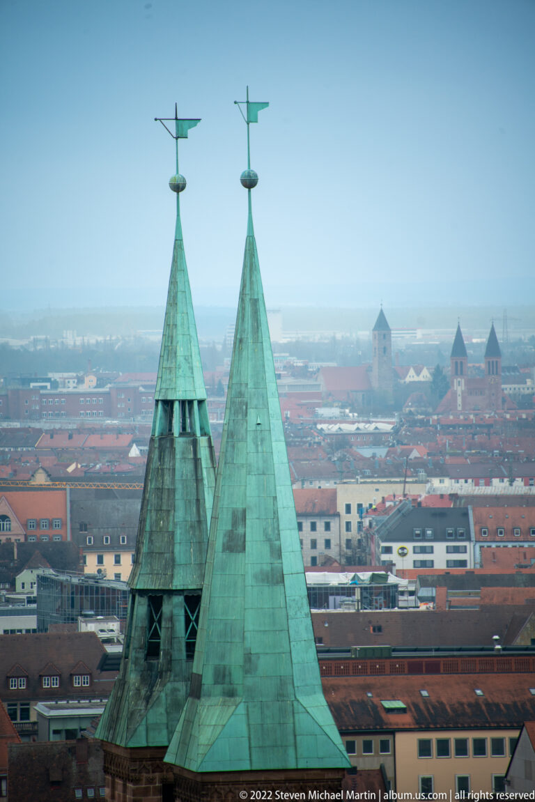 Close elevated view of St. Sebald’s twin green copper spires with weather vanes above Nuremberg rooftops.