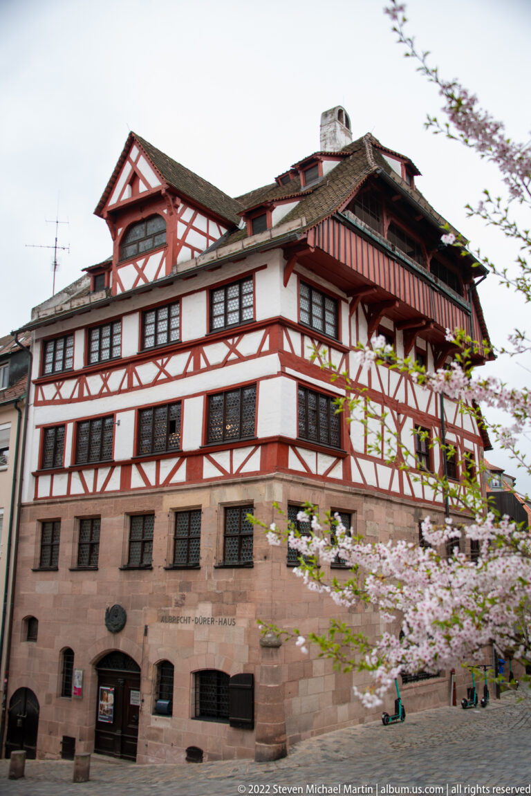Large red-and-white half-timbered building above a sandstone base, with rows of small-paned windows and spring blossoms.