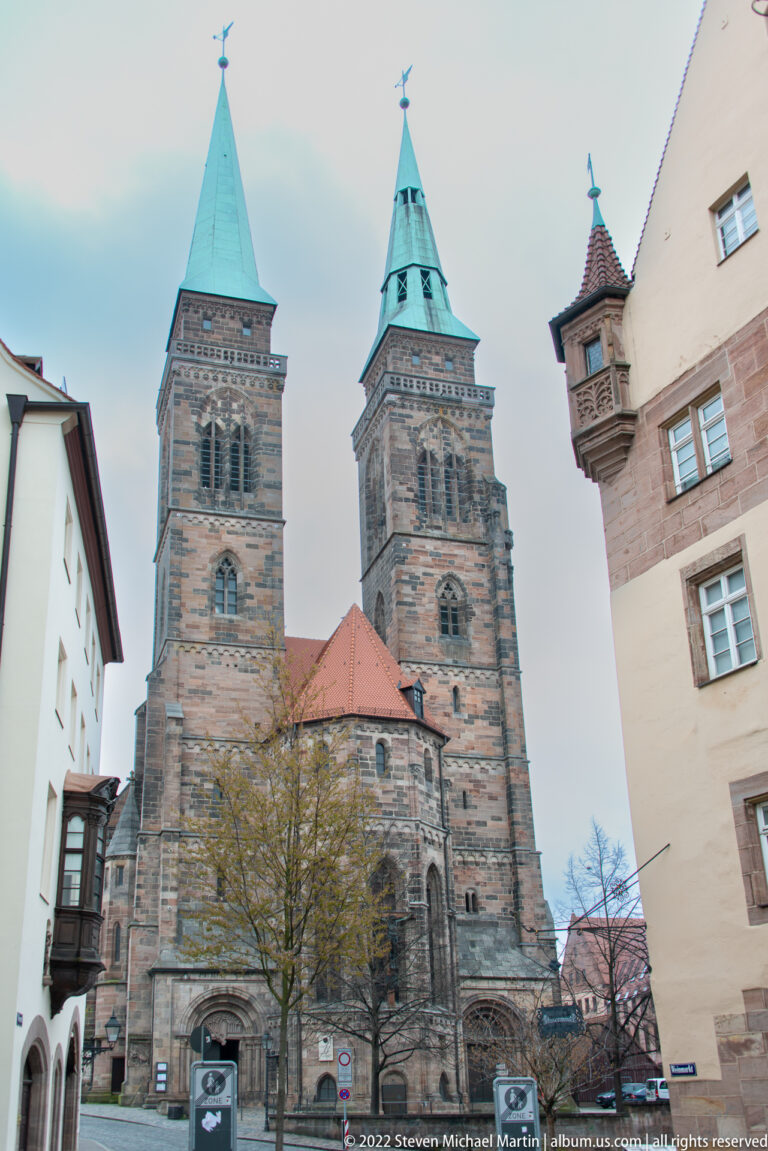Twin green-roofed towers and dark sandstone west front of St. Sebald Church between Old Town buildings.