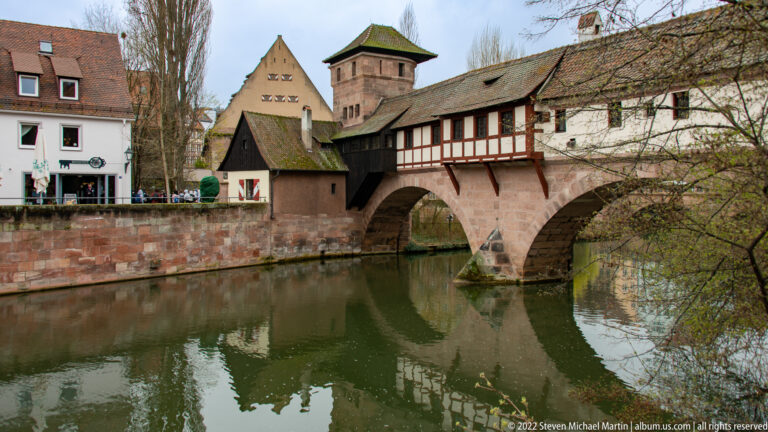 Covered timber bridge on a stone arch crossing the Pegnitz beside historic riverside buildings.