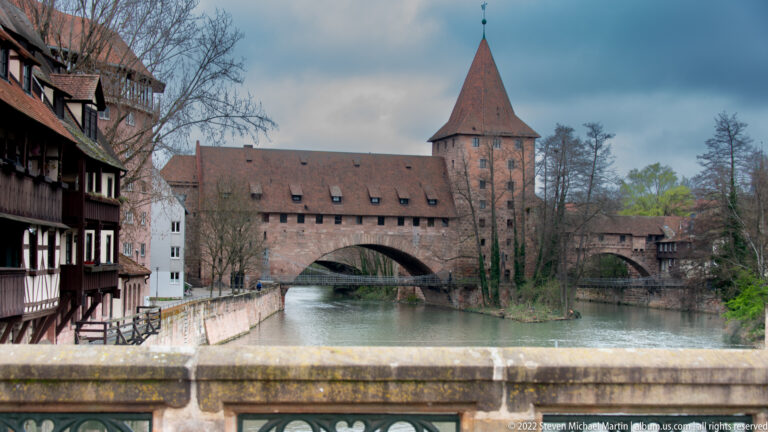 Medieval sandstone tower and roofed bridge spanning the Pegnitz, with river reflections and adjoining historic buildings.