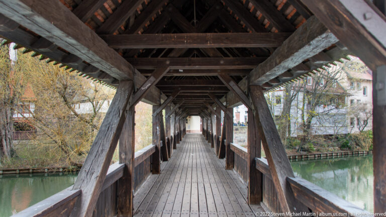 Long covered wooden bridge interior with exposed trusses, plank floor, side rails, and the Pegnitz visible outside.