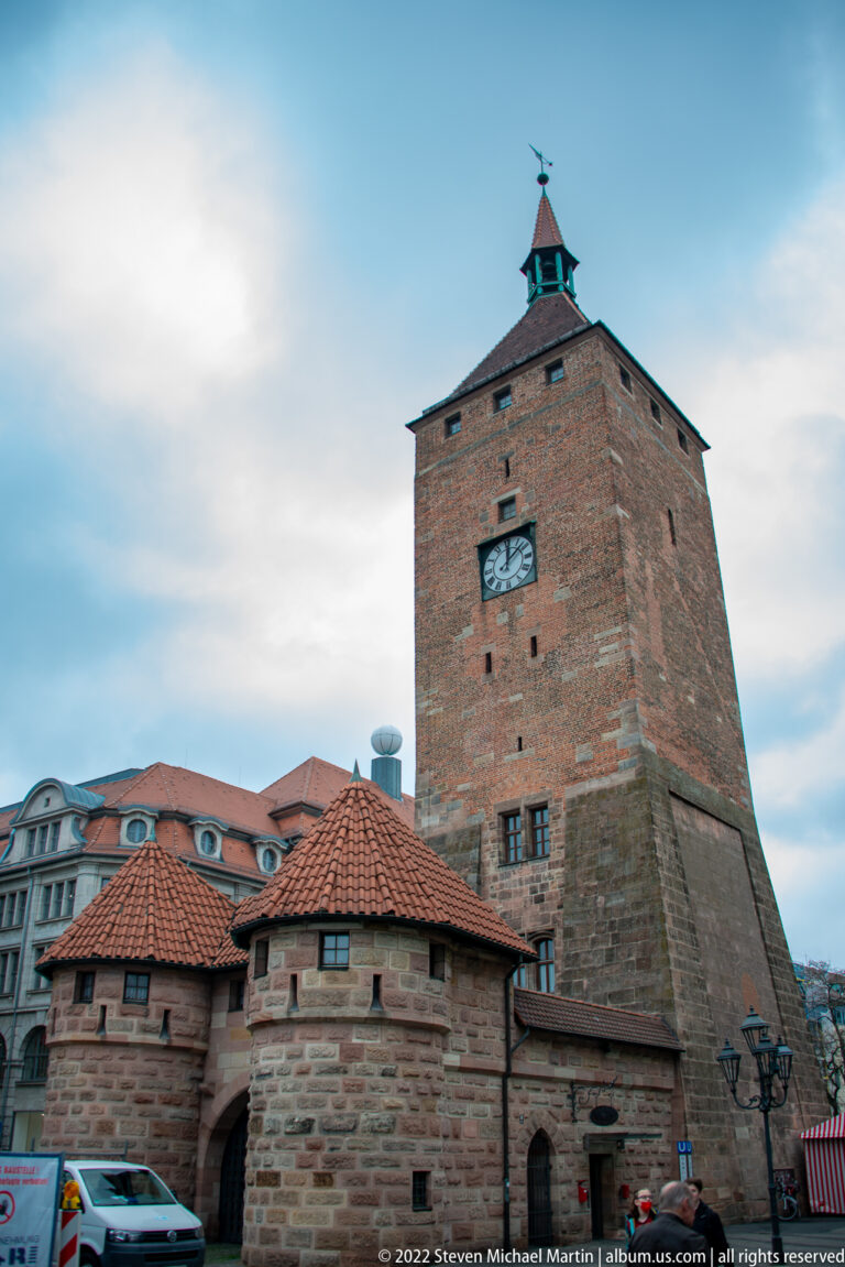 Tall square sandstone gate tower with clock above lower round defensive structures and red tile roofs.