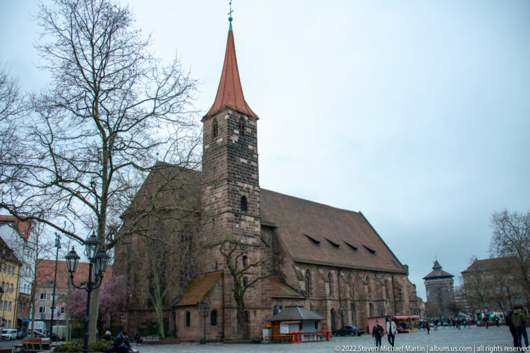 Sandstone Gothic church with steep red roof, tall pointed bell tower, and buttressed side walls.