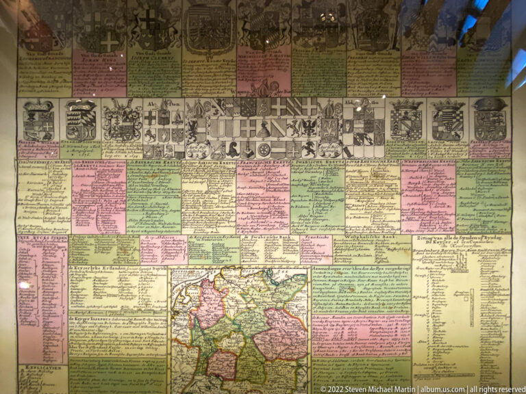 Large historical chart with genealogical text, heraldic shields, portrait medallions, and a colored map at bottom center.
