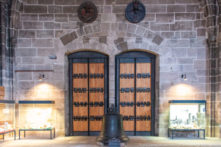 Large dark church bell standing on the floor before paired timber doors and illuminated display cases.