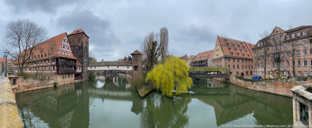 Pegnitz river channel with tree-covered island, historic riverside buildings, bridges, and stone embankments.