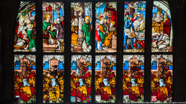 Multi-panel stained-glass window with saints, biblical scenes, heraldic shields, and dark stone mullions.
