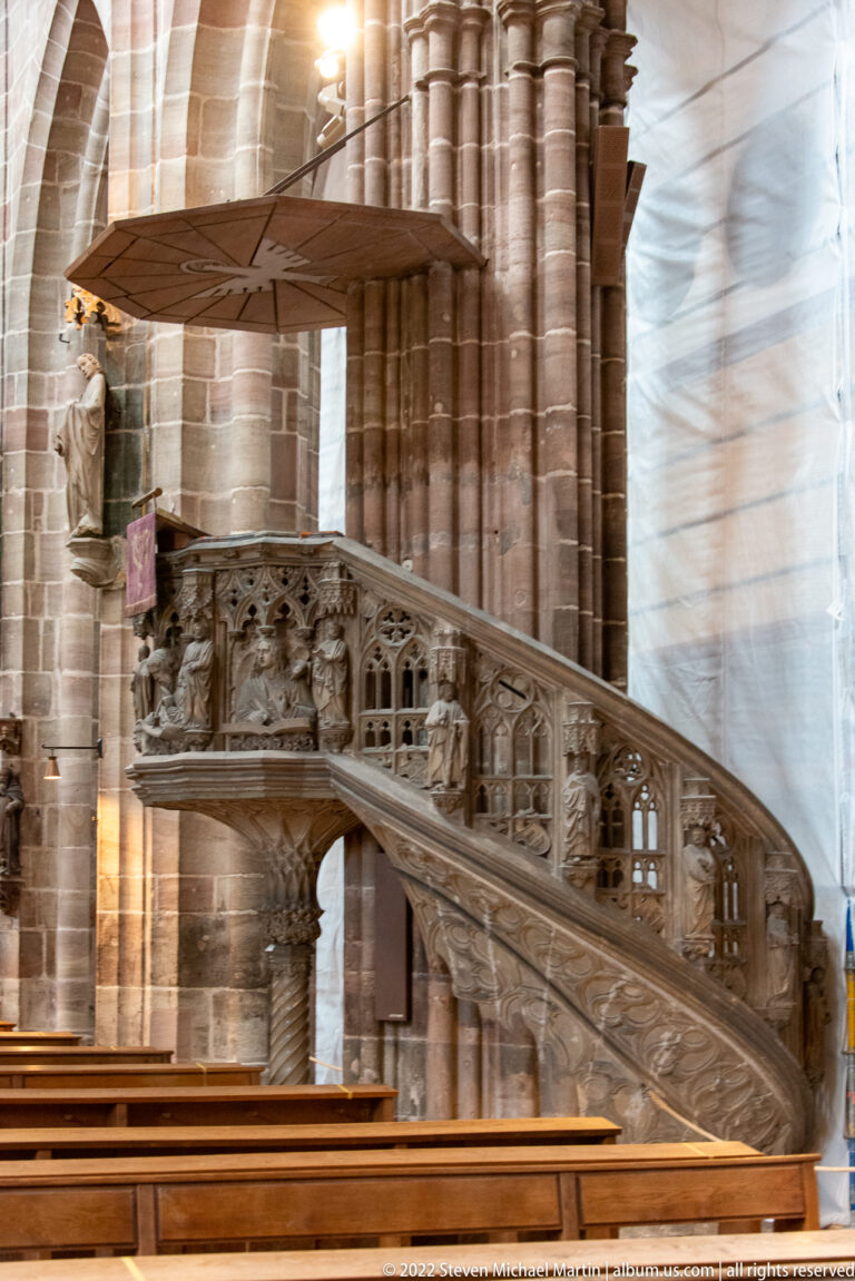 Carved stone pulpit with curving stair, tracery balustrade, and sculpted figures beside massive church columns.
