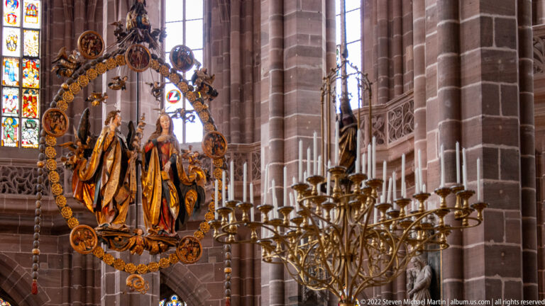 Suspended carved Annunciation group within a circular wreath beside sandstone piers and a large brass candelabrum.