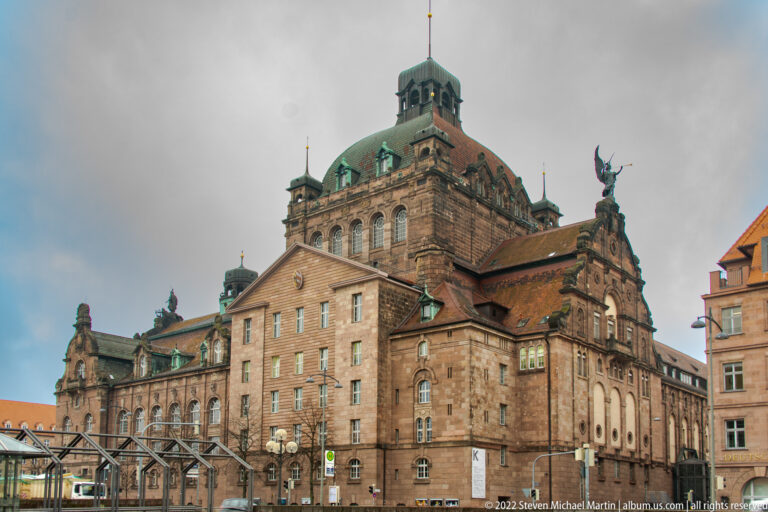 Large sandstone Nuremberg State Theatre building with copper roofs, corner pavilions, and ornate roofline sculpture.