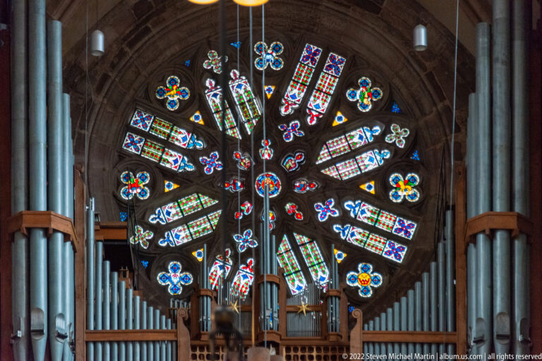Large circular Gothic rose window with radiating colored glass framed on both sides by tall organ pipes.