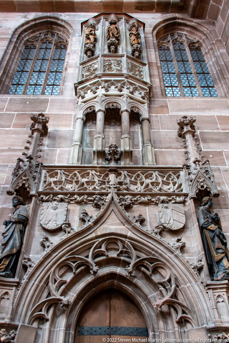 Pointed stone opening surrounded by dense Gothic tracery, coats of arms, figures, and carved architectural ornament.