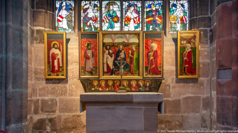 Small painted polyptych with standing saints, a central sacred scene, predella figures, and stained glass above.