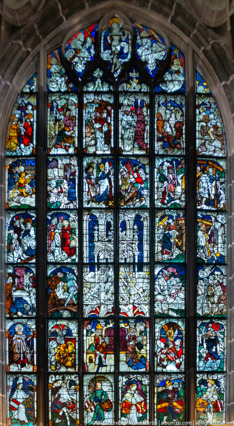 Broad stained-glass window filled with many colorful rectangular narrative panels beneath a curved stone arch.