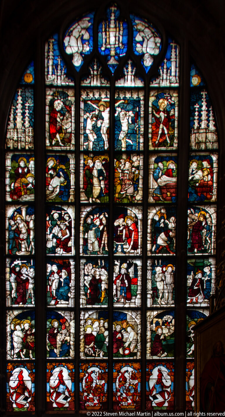 Tall stained-glass window with many stacked figurative scenes divided by dark vertical mullions.