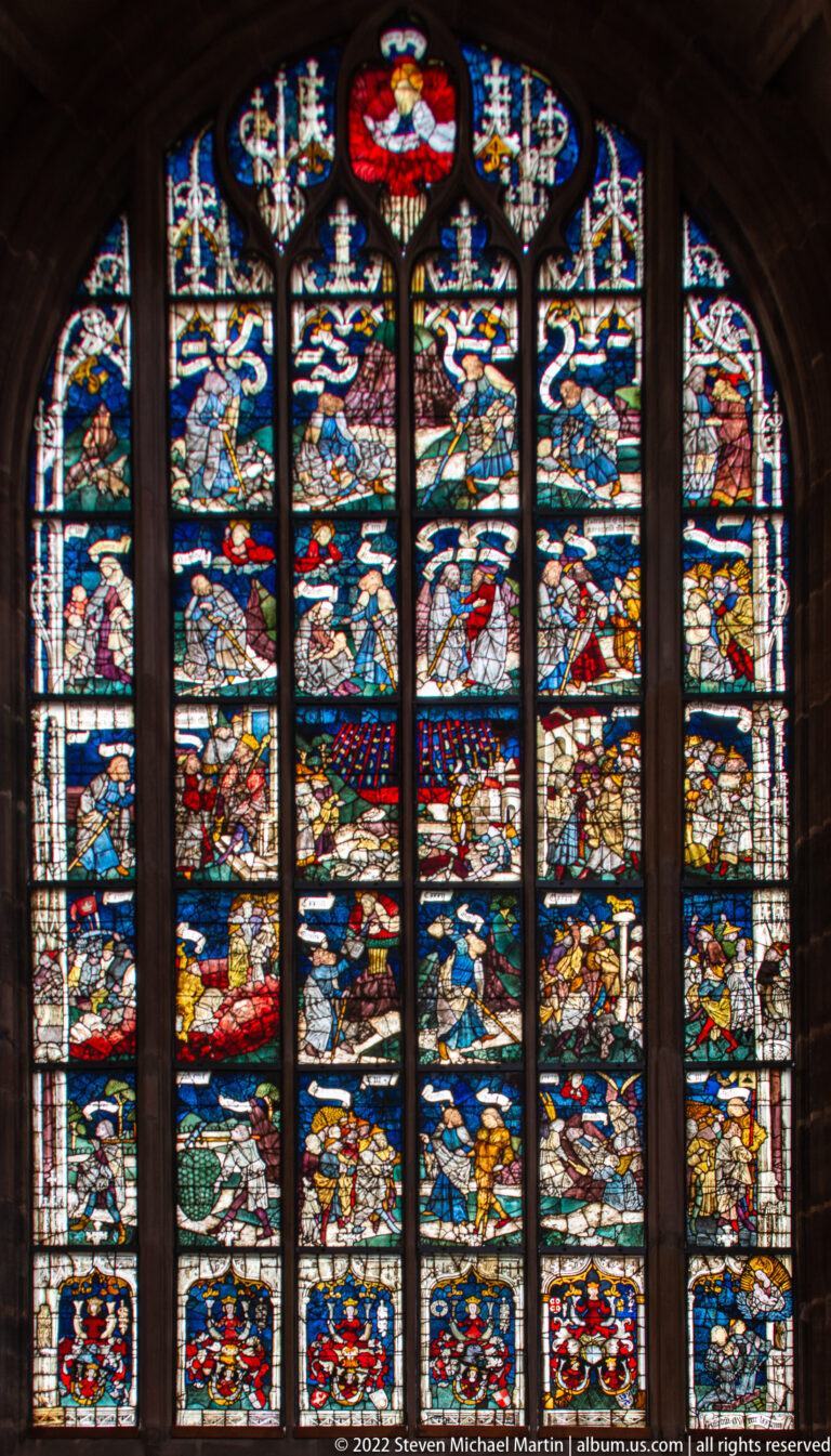 Large multi-lancet stained-glass window with colorful narrative scenes, coats of arms, and Gothic tracery.